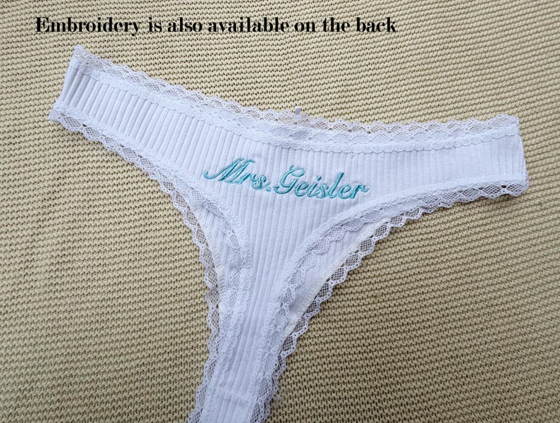 May include: A white ribbed thong featuring delicate lace trim along the edges and the text Mrs. Geisler embroidered in light blue thread on the front. The text Embroidery is also available on the back is visible above the garment.