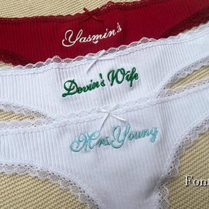 May include: Three pairs of ribbed underwear with lace trim and small bows. The top pair is dark red with white embroidery reading Yasmin's. The middle pair is white with green embroidery reading Devin's Wife. The bottom pair is white with light blue embroidery reading Mrs. Young.