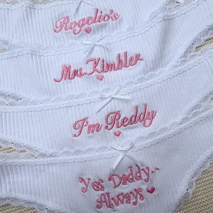 May include: Four pairs of white ribbed underwear featuring lace trim and small white bows. Each pair is embroidered with pink text: Rogelio's, Mrs. Kimbler, I'm Reddy, and Yes Daddy Always, with small pink heart accents included in the designs.