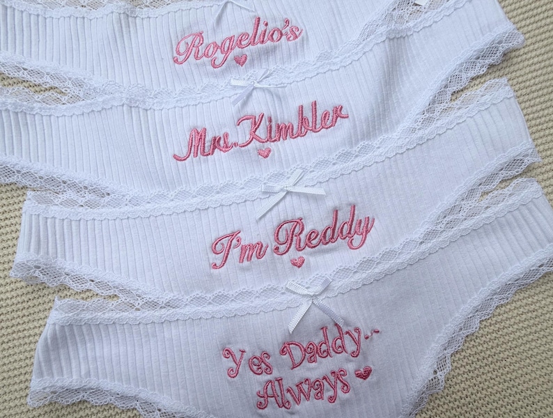 May include: Four pairs of white ribbed underwear featuring lace trim and small white bows. Each pair is embroidered with pink text: Rogelio's, Mrs. Kimbler, I'm Reddy, and Yes Daddy Always, with small pink heart accents included in the designs.