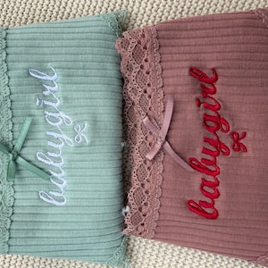 May include: Two ribbed fabric rectangles, one sage green and one dusty rose, each featuring lace trim along one edge, a small satin ribbon bow, and the embroidered text "babygirl" in white and red thread respectively.