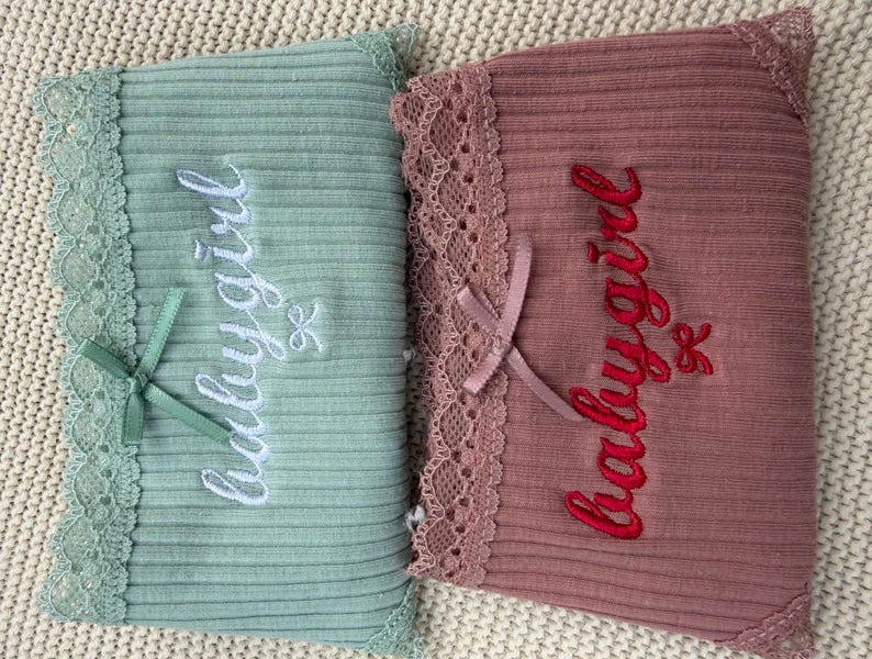 May include: Two ribbed fabric rectangles, one sage green and one dusty rose, each featuring lace trim along one edge, a small satin ribbon bow, and the embroidered text "babygirl" in white and red thread respectively.