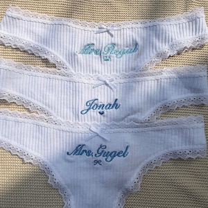 May include: Three white ribbed underwear pieces featuring delicate lace trim along the edges and a small white bow at the center. Each pair is embroidered with blue text reading Mrs. Regal, Jonah, and Mrs. Gugel, accompanied by small icons.
