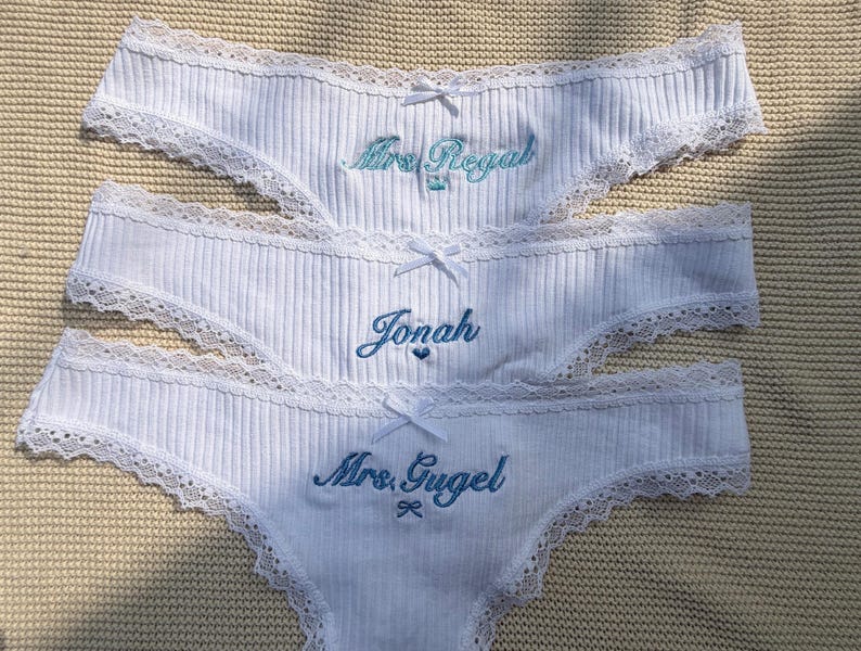 May include: Three white ribbed underwear pieces featuring delicate lace trim along the edges and a small white bow at the center. Each pair is embroidered with blue text reading Mrs. Regal, Jonah, and Mrs. Gugel, accompanied by small icons.