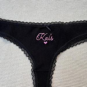 May include: Black ribbed underwear featuring a delicate black lace trim along the edges and a small black bow at the center waist. The front is embroidered with the pink text Kai's above a small pink heart symbol, creating a distinct contrast.
