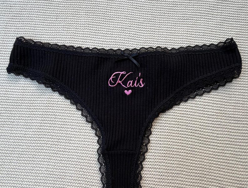 May include: Black ribbed underwear featuring a delicate black lace trim along the edges and a small black bow at the center waist. The front is embroidered with the pink text Kai's above a small pink heart symbol, creating a distinct contrast.
