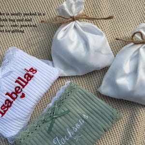 May include: Two ribbed fabric items with lace trim, one white with red embroidered text reading Isabella's and one sage green with white embroidered text reading Jackson's, accompanied by two white satin drawstring pouches tied with brown twine.
