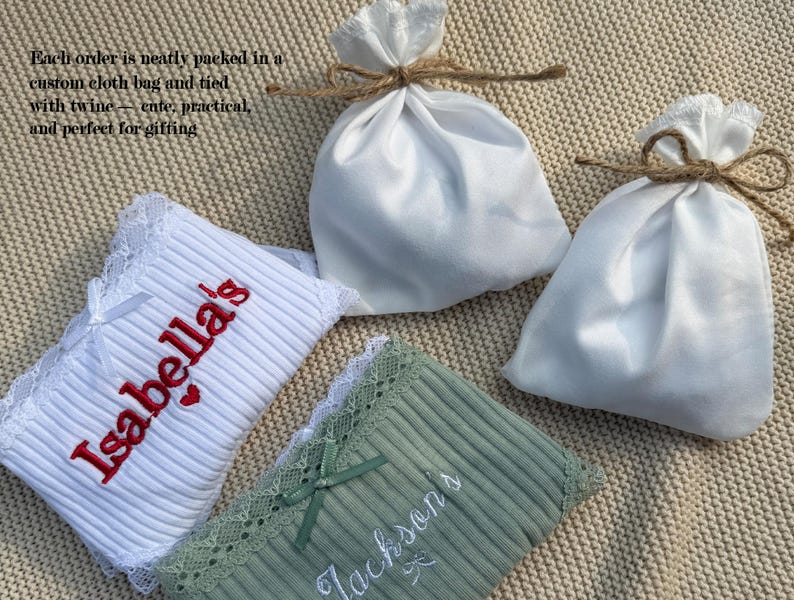 May include: Two ribbed fabric items with lace trim, one white with red embroidered text reading Isabella's and one sage green with white embroidered text reading Jackson's, accompanied by two white satin drawstring pouches tied with brown twine.