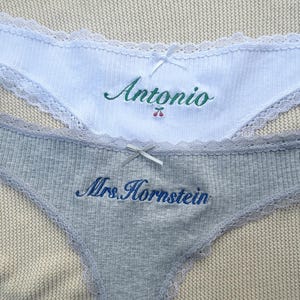 May include: A white ribbed pair of underwear with lace trim, a small white bow, and green embroidered text reading Antonio with a cherry icon. A grey ribbed pair of underwear with lace trim, a grey bow, and blue embroidered text reading Mrs. Hornstein.
