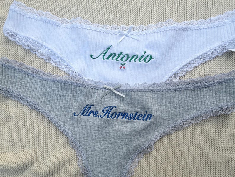 May include: A white ribbed pair of underwear with lace trim, a small white bow, and green embroidered text reading Antonio with a cherry icon. A grey ribbed pair of underwear with lace trim, a grey bow, and blue embroidered text reading Mrs. Hornstein.