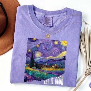 May include: A violet short-sleeved t-shirt featuring a graphic print of a starry night landscape with swirling clouds, a dark cypress tree, and a village. The text "LET IT GOGH" is printed in white below the graphic. The shirt has a Comfort Colors label.