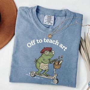 May include: A blue t-shirt featuring a graphic of a green frog wearing a maroon beret, round glasses, and an apron, riding a scooter. The frog holds a basket containing paintbrushes. The text "Off to teach art" is printed in white above the frog.