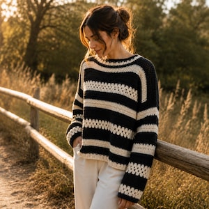 May include: A black and cream striped crochet sweater with a round neckline and long sleeves. The sweater is worn with white pants. The person is leaning against a wooden fence in a field.