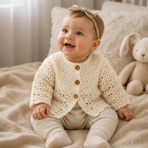 May include: A cream-coloured, long-sleeved crochet cardigan featuring a round neckline, three small brown buttons down the front, and a scalloped hem. The garment is worn over light beige leggings, paired with a matching cream-coloured bow headband.