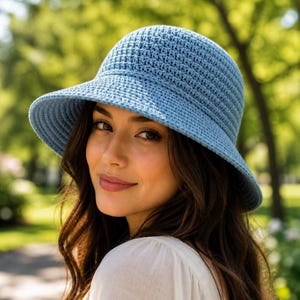 May include: A light blue crochet bucket hat with a rounded crown and a downward-sloping brim. The hat is constructed from a dense, uniform stitch pattern in a solid, muted blue yarn, providing a structured yet flexible shape for the head.