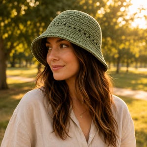 Puede incluir: Un sombrero de cubo de ganchillo verde salvia con un ala ligeramente curvada. El sombrero tiene una apariencia texturizada y una serie de pequeños agujeros espaciados uniformemente cerca de la parte superior. El sombrero lo lleva una persona con cabello castaño largo y ondulado.