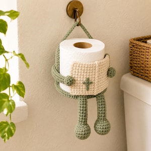 Könnte beinhalten: Ein salbeigrüner gehäkelter Toilettenpapierhalter in Form einer Figur, deren Arme eine Rolle weißes Toilettenpapier umschließen. Die Figur hält ein kleines cremefarbenes gehäkeltes Buch mit einem grünen Kreuz auf dem Einband.