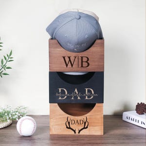 Op de afbeelding: Een verticale stapel van drie vierkante houten opbergdozen voor honkbalpetten. De bovenste doos is donkerbruin met de letters WB. De middelste doos is zwart met de tekst DAD en namen. De onderste doos is lichtbruin met DAD en gewei-afbeeldingen.