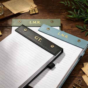 Op de afbeelding: Een close-up van een notitieblok met een zwarte leren kaft en gelijnd papier. Drie leren penhouders in zwart, groen en blauw zijn bevestigd, elk met gouden initialen. Gouden paperclips en vintage papier zijn verspreid.