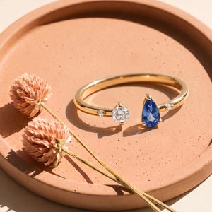 Puede incluir: Un anillo abierto de oro con un zafiro azul en forma de pera y un diamante redondo de talla brillante, acentuado por dos diamantes redondos más pequeños en la banda. El anillo se muestra junto a dos flores secas de amaranto rosa claro.
