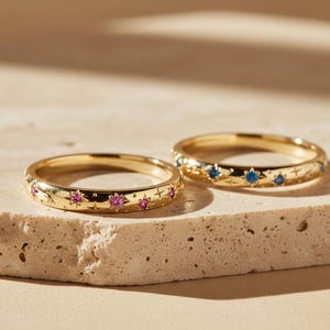 Op de afbeelding: Twee gouden ringen met stervormige gravures en kleine ingelegde edelstenen. De ene ring is versierd met roze stenen, terwijl de andere blauwe stenen heeft. Beide ringen hebben een gepolijste afwerking met geëtste sterpatronen rondom elke steen.