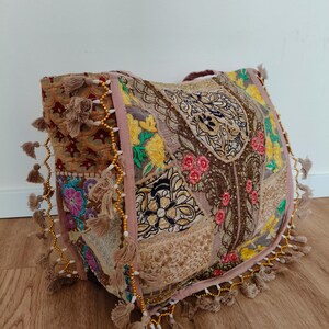 Könnte beinhalten: Eine große Tragetasche mit einem Patchwork-Design aus bestickten Stoffen in Rosa-, Gold-, Gelb- und Brauntönen. Die Tasche ist mit zahlreichen kleinen beigen Quasten und goldfarbenen Perlenketten an den Kanten und Seitenteilen verziert.