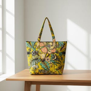 Pode incluir: Uma bolsa tote acolchoada com uma base amarelo-mostarda vibrante e um padrão floral multicolorido em tons de azul-petróleo, pêssego e cinza-escuro. A bolsa tem duas alças de tecido a condizer e pequenas abas laterais pretas com rebites de metal prateado.