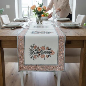 Könnte beinhalten: Ein rechteckiger, cremeweißer Tischläufer mit einem sich wiederholenden floralen Blockdruckmuster in gedämpftem Orange und Dunkelgrau. Der Läufer hat eine dekorative Bordüre mit geometrischem Design und liegt auf einem hölzernen Esstisch.