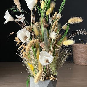 Puede incluir: Un arreglo floral seco con amapolas blancas, trigo y hierbas en un jarrón geométrico de hormigón gris. El arreglo incluye elementos verdes, amarillos y beige, creando una exhibición natural y texturizada.