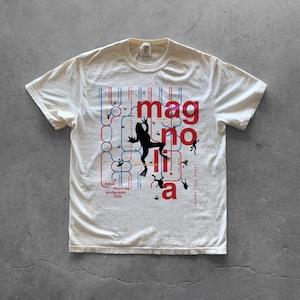 Könnte beinhalten: Ein cremefarbenes Kurzarm-T-Shirt mit einem Grafikdruck, der das Wort Magnolia in großen roten Buchstaben zeigt. Das Design enthält eine schwarze Froschsilhouette, blaue und rote Liniendiagramme sowie den Text a Paul Thomas Anderson film.
