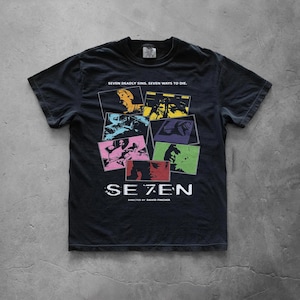 Può includere: Una t-shirt nera a maniche corte con una stampa grafica che riporta il testo SE7EN, DIRECTED BY DAVID FINCHER, e SEVEN DEADLY SINS. SEVEN WAYS TO DIE. Il design include un collage di cornici rettangolari colorate con varie scene di film.