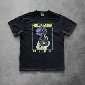 May include: Black t-shirt featuring a graphic from the movie "Hellraiser: Bloodline." The design includes the film's title, a hand holding a puzzle box, and a purple and blue visual element. The shirt has a crew neck and short sleeves.