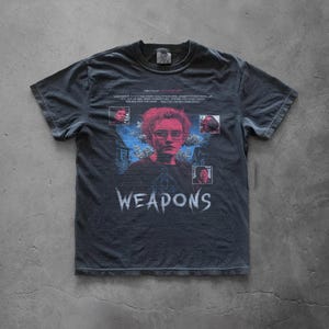 Peut inclure: Un t-shirt gris anthracite à manches courtes avec une impression graphique représentant un portrait central en tons roses, entouré de petites images encastrées, de silhouettes d'arbres bleues et du texte blanc WEAPONS sur la poitrine.