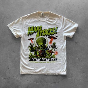 May include: An off-white short-sleeve t-shirt featuring a graphic print of a green alien from Mars Attacks. The design includes the text MARS ATTACKS!, ACK! ACK! ACK!, and NICE PLANET, WE'LL TAKE IT!, along with flying saucers and a ray gun.