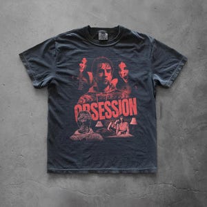 Könnte beinhalten: Ein anthrazitfarbenes Kurzarm-T-Shirt mit rotem Grafikdruck. Das Design zeigt ein zentrales Porträt, kleinere Figuren und das Wort OBSESSION in fetten roten Buchstaben. Der Text lautet: BE CAREFUL WHO YOU MESS WITH.