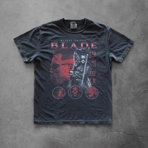 May include: Dark gray t-shirt featuring a graphic of Wesley Snipes as Blade. The design includes the word "BLADE" in red, a portrait of Snipes, and other graphic elements. The shirt has a vintage, worn look.