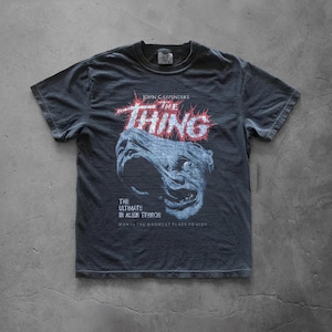 Peut inclure: T-shirt gris foncé avec le titre du film "The Thing" en rouge et blanc, et un dessin de monstre. Le texte "The Ultimate in Alien Terror" et "Man is the warmest place to hide" sont également affichés.