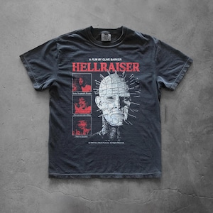 May include: A charcoal gray short-sleeve t-shirt featuring a graphic print from the film Hellraiser. The design includes the text A FILM BY CLIVE BARKER, HELLRAISER, three small red-toned character panels, and a large white illustration of Pinhead.