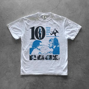 Könnte beinhalten: Weißes T-Shirt mit einem blauen Aufdruck aus dem Film "10 Dinge, die ich an dir hasse". Das Design zeigt die Zahl 10, Silhouetten von Charakteren und den Filmtitel. Darunter befinden sich Bilder der Schauspieler des Films.