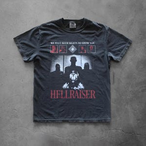 May include: A charcoal gray short-sleeve t-shirt featuring a graphic print from the movie Hellraiser. The design includes the text WE HAVE SUCH SIGHTS TO SHOW YOU, four small character portraits, a central figure, and the red text HELLRAISER.