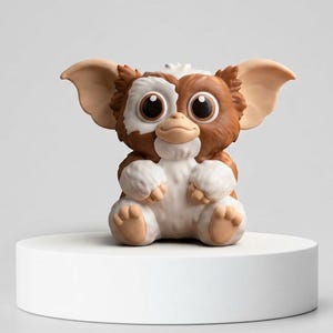 Puede incluir: Una pequeña figura estilizada de una criatura con orejas grandes y puntiagudas, un patrón de pelaje marrón y blanco y ojos oscuros de gran tamaño. La figura está representada en posición sentada con pelaje blanco en el pecho, las patas y los pies, y tiene un acabado mate y suave.