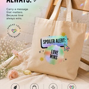 Op de afbeelding: Een beige canvas draagtas met een grafische print met de tekst "SPOILER ALERT:" in een zwart kader en "LOVE WINS" in een regenboogkleurige wolkvorm, inclusief een computercursor-icoon en kleine steraccenten op de stof.