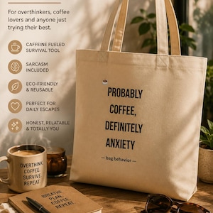 May include: A beige canvas tote bag featuring the black printed text PROBABLY COFFEE, DEFINITELY ANXIETY and - bag behavior - with a small coffee cup icon above the text. The bag has two long shoulder straps and a rectangular structure.