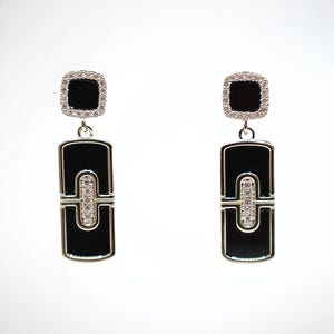 Puede incluir: Un par de pendientes elegantes con una piedra cuadrada de ónix negro rodeada de pequeños cristales brillantes. Los pendientes tienen una caída rectangular de ónix negro con un acento central de cristal, suspendido de una parte superior cuadrada más pequeña.