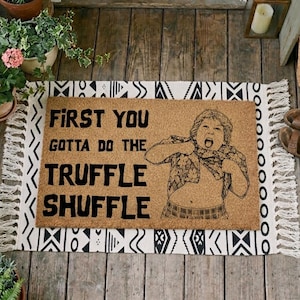 Könnte beinhalten: Eine rechteckige Fußmatte mit dem Text "FIRST YOU GOTTA DO THE TRUFFLE SHUFFLE" in Schwarz. Die Matte zeigt eine Grafik eines Kindes und ist von einem schwarz-weiß gemusterten Teppich mit Fransen umgeben.