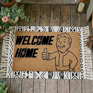 May include: A rectangular doormat with the words "WELCOME HOME" in black, and a cartoon character giving a thumbs-up. The mat is light brown and is bordered by a black and white patterned rug. The doormat is suitable for a front door.