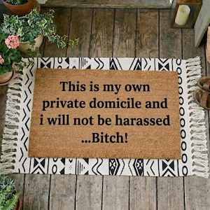 Pode incluir: Um capacho retangular com o texto "This is my own private domicile and I will not be harassed...Bitch!" em preto. O tapete é castanho com uma borda e franjas estampadas em preto e branco.