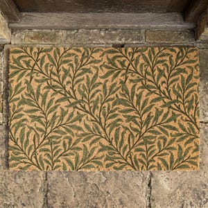 Puede incluir: Felpudo rectangular con fondo beige y un patrón repetido de hojas y vides verdes. El diseño simétrico cubre toda la superficie de la alfombra, creando un tema natural y botánico.