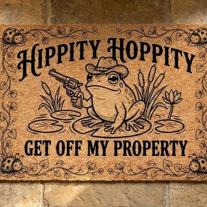 Op de afbeelding: Kokosmat met de tekst "Hippity Hoppity Get Off My Property". De mat toont een cartoonkikker met een cowboyhoed en een revolver. Decoratieve bloemen- en lieveheersbeestjesaccenten omlijsten de tekst en de kikker.