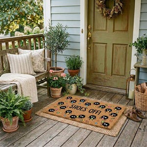 May include: A welcoming front porch scene with a wooden bench, potted plants, and a doormat. The doormat is brown with a repeating cartoon character design and the words "SHOES OFF!". A wreath hangs on the green front door. A pair of brown shoes sits near the mat.
